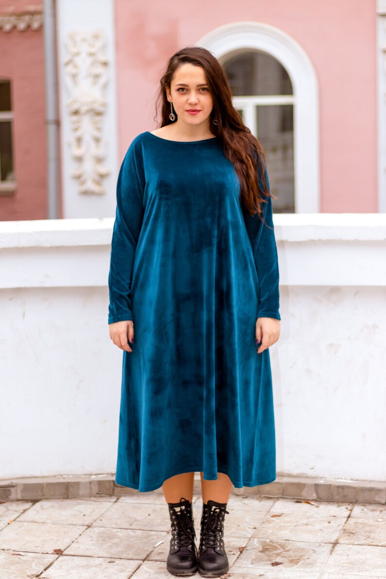 May include: A teal blue velvet midi dress with long sleeves and a round neckline. The dress is loose fitting and falls to the knees. The model is wearing black combat boots.