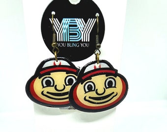 May include: A pair of earrings featuring a cartoon character with a red hat and a big smile. The earrings are made of metal and have a gold finish. The earrings are hanging from a black card with the text "You Bling You" printed on it.
