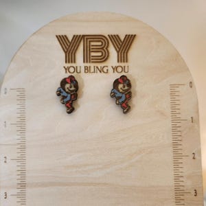 May include: A pair of wooden earrings featuring a cartoon character wearing a red hat and blue shirt. The earrings are displayed on a wooden surface with a ruler for scale. The text "YBY You Bling You" is visible on the surface.