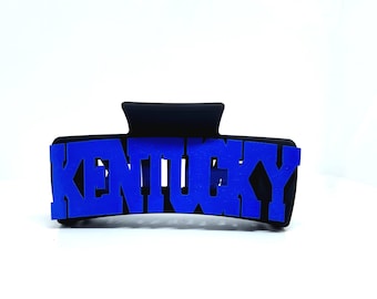 May include: Black hair claw clip with the word "KENTUCKY" in blue block letters. The clip is a rectangular shape with a curved top and a hinged mechanism for securing hair. The letters are bold and easy to read.