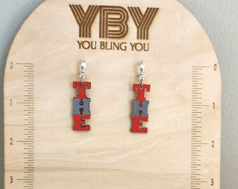 May include: A pair of red and gray wooden earrings spelling out the word "THE" are displayed on a wooden stand. The earrings hang from silver-colored hoops. The stand has the text "YBY YOU BLING YOU" engraved on it.
