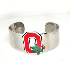 May include: A silver metal bracelet with a red and white letter "O" charm with a green leaf and a small red berry.