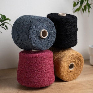 Può includere: Quattro rocchetti di filato blu, nero, bordeaux e oro sono impilati su una superficie di legno. Il filato ha un aspetto strutturato e ad anello. Ogni rocchetto ha un foro centrale con un'anima di cartone chiaro.