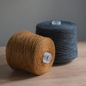 Puede incluir: Dos bobinas de hilo, una en marrón cálido y otra en gris oscuro, sobre una superficie de madera. El hilo está bien enrollado alrededor de un núcleo de plástico transparente. El fondo es gris liso.