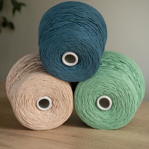 Può includere: Tre rocchetti di filo color verde acqua, verde chiaro e beige sono impilati. Ogni rocchetto ha un centro bianco. Il filo è ben avvolto e i colori sono tenui e invitanti. I rocchetti sono disposti su una superficie di legno.