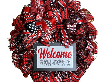 May include: Red, black, and white wreath with a sign that says "Welcome" in sign language. The wreath is made of mesh and ribbon.