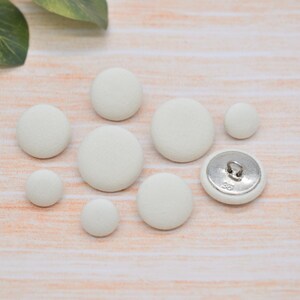 May include: An assortment of off-white fabric-covered buttons in different sizes, one with a silver metal shank. The buttons are displayed on a light-coloured wooden surface, with green leaves in the background.