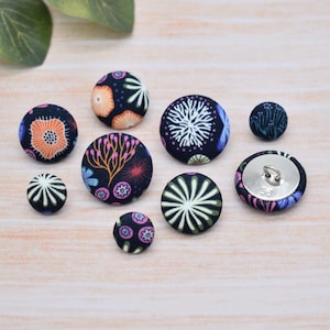 May include: A selection of fabric-covered buttons in different sizes. The buttons display floral and abstract patterns in orange, pink, white, and green on a navy blue backdrop. Some have a silver metal shank.