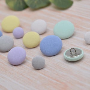 May include: Assortment of fabric covered buttons in various pastel colours, including light blue, lavender, yellow, and grey. The buttons are arranged on a light wood surface.