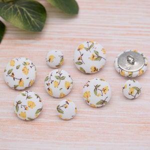 May include: Assortment of eight fabric covered buttons with a floral pattern. The buttons are white with yellow flowers and brown stems. One button has a silver metal back.