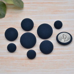 May include: A selection of navy blue fabric-covered buttons in different sizes. The buttons are displayed on a light wooden surface, with some green leaves in the top left. One button reveals its metal backing.
