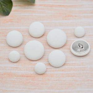 May include: A selection of white fabric-covered buttons in different sizes, one with a metal shank. The buttons are displayed on a light-coloured wooden surface, with green leaves in the top left corner.