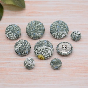 May include: Assortment of eight fabric covered buttons in various shades of green and blue with a white background. The buttons feature a floral pattern and are arranged on a light wood surface.