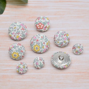 Op de afbeelding: Assortiment van acht met stof beklede knoppen met een bloemenpatroon in roze, gele en groene tinten op een witte achtergrond. De knoppen zijn in verschillende maten.