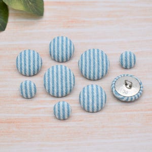 Pode incluir: Uma coleção de botões redondos revestidos de tecido em vários tamanhos. Os botões apresentam um padrão de listras azuis claras e brancas. Um botão mostra a parte traseira de metal com o número 38. Os botões estão dispostos em uma superfície clara de grão de madeira.