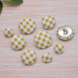 May include: Assortment of eight yellow and white gingham fabric buttons. The buttons are round and have a white background with a yellow gingham pattern.