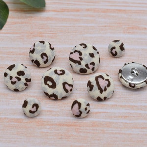 May include: A set of eight fabric covered buttons in various sizes. The buttons feature a white background with a brown and pink leopard print.