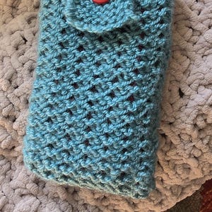 May include: A teal blue, hand-knitted phone case with a flap and a red button closure. The case is made of a textured yarn and is resting on a soft, light grey blanket.