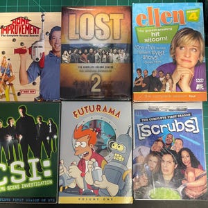 May include: A collection of DVD cases for popular television shows, including Home Improvement, Lost, Ellen, Futurama, Scrubs, and CSI. The cases are arranged in a row on a green surface.