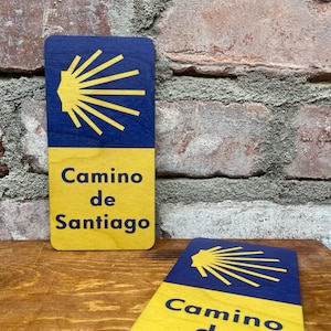 Puede incluir: Dos letreros de madera amarillos y azules con el símbolo de la concha de vieira y el texto "Camino de Santiago".