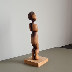 Peut inclure: Sculpture en bois représentant une figure humaine stylisée. La figurine est sculptée dans du bois brun clair et foncé, avec une finition lisse. Elle repose sur une base carrée en bois. Design minimaliste et abstrait.