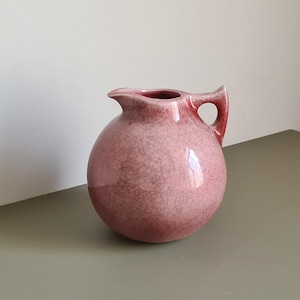 Peut inclure: Pichet rond en céramique rose avec une anse et un bec verseur. Le pichet présente une finition craquelée, lui donnant un aspect texturé. La poignée comporte une ouverture circulaire. Le pichet est probablement utilisé pour servir des boissons ou comme objet décoratif.