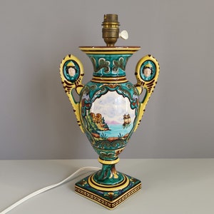 May include: A green and yellow ceramic table lamp shaped like an urn with a painted scene of a coastal landscape and two figures on the handles.