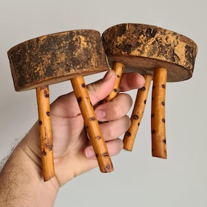 Peut inclure: Deux petits tabourets en bois faits à la main. Chaque tabouret a une assise ronde en bois naturel et trois pieds fins brun clair avec des taches sombres. Les assises ont un aspect texturé semblable à de l'écorce.