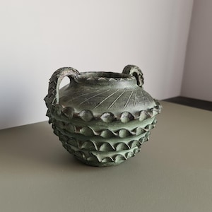 Può includere: Un piccolo recipiente rotondo verde scuro con due manici arcuati. L'esterno presenta una superficie testurizzata con tre file orizzontali di creste smerlate a forma di onda e linee radianti attorno all'apertura superiore.