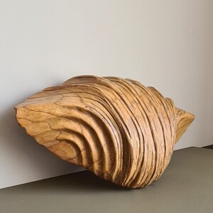 May include: A large, carved wooden seashell sculpture. The sculpture is a light brown color and has a textured surface with detailed ridges and curves. The seashell is positioned on a neutral background.