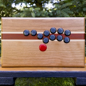 Op de afbeelding: Een houten arcade-bedieningspaneel met zwarte knoppen en een rode knop in het midden. Het paneel heeft een kersenhouten afwerking met een rode en witte streep.