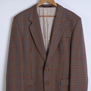 Può includere: Un blazer a quadri marrone, blu e rosso dal design classico. Il blazer ha un revers a lancia, tre bottoni e due tasche anteriori. È appeso a una gruccia di legno su sfondo bianco.