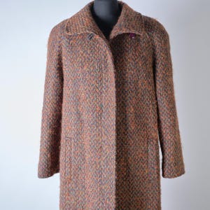Può includere: Un cappotto vintage al ginocchio in un tessuto strutturato e maculato. Il cappotto presenta un colletto a revers, maniche lunghe e una chiusura con un solo bottone. La tavolozza dei colori include sfumature di marrone, arancione e blu.