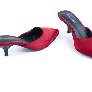 May include: A pair of red satin heeled mules with a pointed toe. The shoes have a black leather insole and a black heel. The brand name "Jeffrey Campbell" is printed on the insole.