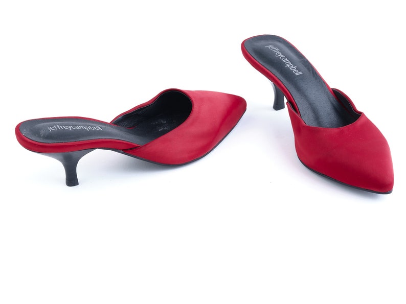 May include: A pair of red satin heeled mules with a pointed toe. The shoes have a black leather insole and a black heel. The brand name "Jeffrey Campbell" is printed on the insole.