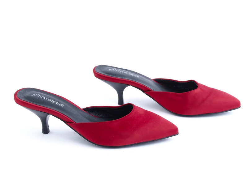May include: A pair of red satin heeled mules with a pointed toe. The shoes have a black heel and the brand name "jeffreycampbell" is printed on the insole.