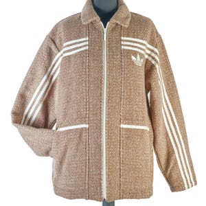May include: A brown zip-up jacket with white stripes on the sleeves and a white collar. The jacket features a white zipper and two front pockets. The Adidas logo is visible on the chest.