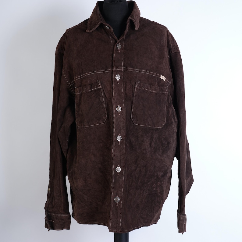 Puede incluir: Una camisa de ante marr&oacute;n con cierre de botones y dos bolsillos grandes en el pecho. La camisa tiene una peque&ntilde;a etiqueta en el bolsillo derecho que dice "Buffalo".