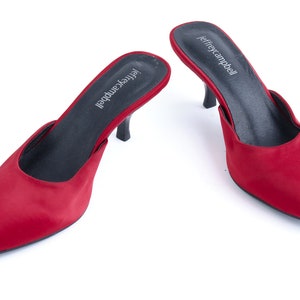 May include: A pair of red satin heeled mules with a pointed toe. The shoes have a black insole and a black heel. The brand name "jeffreycampbell" is printed on the insole.