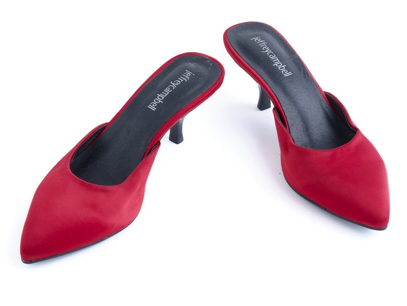 May include: A pair of red satin heeled mules with a pointed toe. The shoes have a black insole and a black heel. The brand name "jeffreycampbell" is printed on the insole.