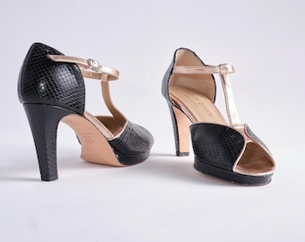 Puede incluir: Un par de zapatos de tacón negros con estampado de piel de serpiente. Los zapatos tienen un diseño de correa en T con una correa y ribete dorados. Los tacones miden aproximadamente 10 cm de alto.
