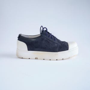 May include: A navy blue suede shoe with a white toe cap and a thick white sole. The shoe has navy blue laces and a black leather trim around the ankle. The sole is approximately 2 inches thick.
