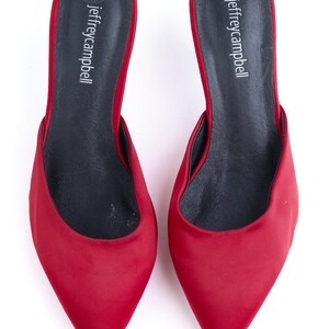May include: A pair of red satin heeled mules with a pointed toe. The shoes have a black leather insole with the brand name "jeffreycampbell" printed on them.
