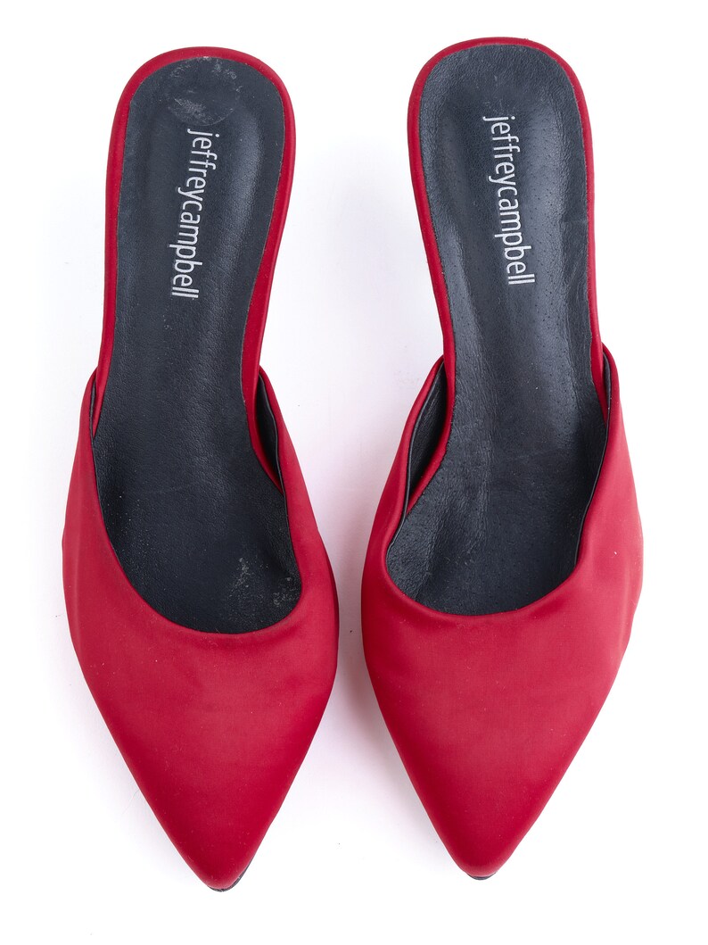 May include: A pair of red satin heeled mules with a pointed toe. The shoes have a black leather insole with the brand name "jeffreycampbell" printed on them.