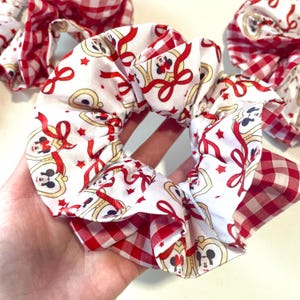 May include: Scrunchies featuring a red and white gingham pattern and a white fabric with a Mickey Mouse and ribbon design. These fabric scrunchies are designed to hold hair in place.
