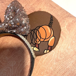 May include: Brown mouse ear headband with a sequined bow. One ear features a cartoon illustration of Slinky Dog from Toy Story. The headband has a gold rope trim and a black base.