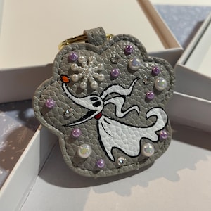 Può includere: Un portachiavi in ​​pelle grigia con un'illustrazione in bianco e nero di Zero, il cane fantasma di The Nightmare Before Christmas. Il portachiavi è decorato con perline bianche e viola e strass.