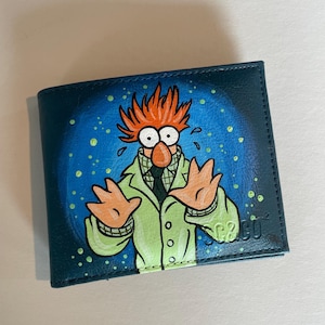 Peut inclure: Un portefeuille bleu foncé à deux volets orné d'une illustration peinte à la main représentant un personnage aux cheveux orange ébouriffés, aux grands yeux blancs et au nez orange rond. Il porte une blouse vert clair sur une chemise à carreaux.