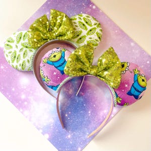 Peut inclure: Bandeaux d'oreilles fantaisie sur le thème de l'espace. Les oreilles présentent un imprimé extraterrestre vert et un fond rose avec un personnage extraterrestre. Les bandeaux ont des nœuds à paillettes vertes et un bandeau en plastique transparent.