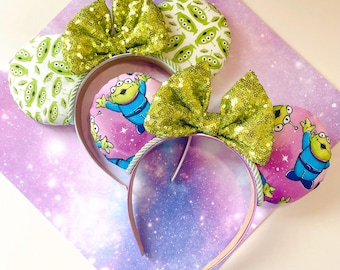 May include: A pair of novelty ear headbands featuring a space theme. The ears have a green alien print and a pink background with an alien character. The headbands have green sequin bows and a clear plastic band.